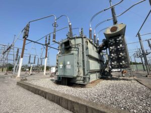 Large high-voltage power transformer with cooling fans at an outdoor electrical substation under clear blue skies.